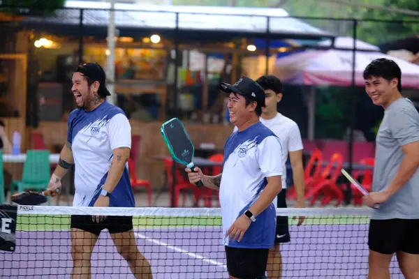 Three men in matching TSK team jerseys laughing together at the net at RC Rally Hub, with a food stall in the background