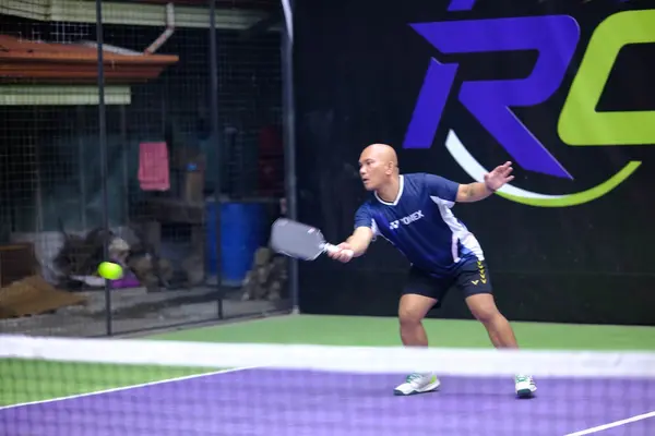 A bald man in a navy Yonex jersey reaching out for a forehand shot, RC Rally Hub banner in the background